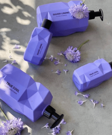 Nine Yards Silver Lining ir True Cool plaukų priežiūros priemonės violetiniuose buteliukuose, išdėliotos su gėlėmis natūralioje aplinkoje