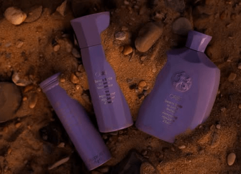 Oribe plaukų priežiūros priemonės violetiniuose buteliukuose ant natūralaus smėlio fono